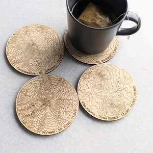 May include: Four wooden coasters with laser-etched topographic maps of mountains. The coasters feature the names and elevations of the mountains:  Seakseekque - Mt. Jefferson - 10,502', Wyeast - Mt. Hood - 11,249', Klah Kiahne - Three Sisters - 10,090', 10,052', 10,363', and Mt. Bachelor - 9,068'.