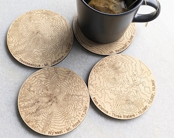 May include: Four wooden coasters with laser-etched topographic maps of mountains. The coasters feature the names and elevations of the mountains:  Seakseekque - Mt. Jefferson - 10,502', Wyeast - Mt. Hood - 11,249', Klah Kiahne - Three Sisters - 10,090', 10,052', 10,363', and Mt. Bachelor - 9,068'.