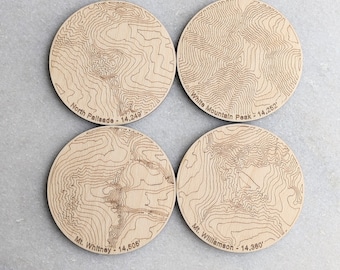 May include: Four wooden coasters with engraved maps of mountains. Each coaster features the name and elevation of a mountain: North Palisade - 14,249', White Mountain Peak - 14,252', Mt. Whitney - 14,505', and Mt. Williamson - 14,380'.
