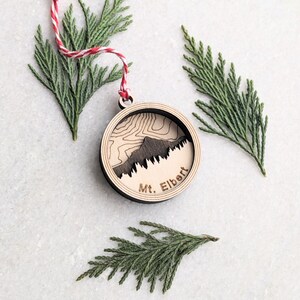 May include: A wooden ornament featuring a laser-cut design of Mount Elbert, a mountain in Colorado. The ornament is round and has a brown wood finish. The text "Mt. Elbert" is engraved on the bottom of the ornament.