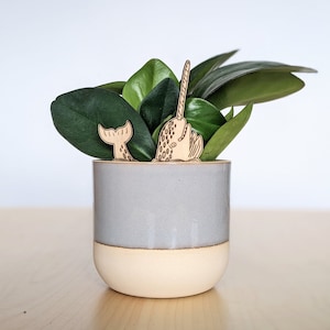May include: A gray ceramic planter with a light brown rim, filled with a green leafy plant. Two wooden narwhal figurines are placed in the planter, one with its tail sticking out and the other with its horn pointing up.