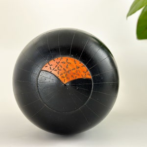 May include: A black wooden sphere with a carved design. The design is a half circle with a pattern of small orange leaves.