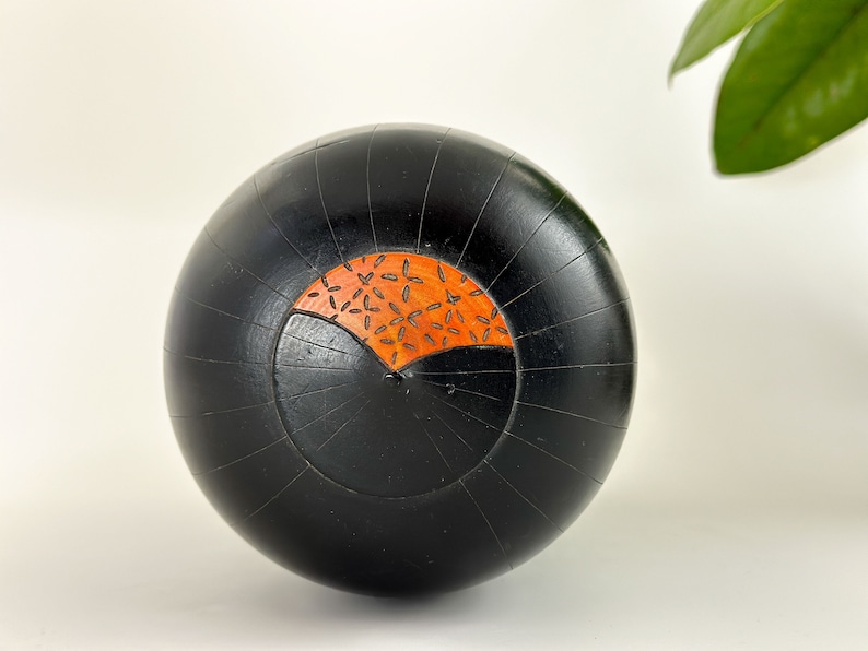 May include: A black wooden sphere with a carved design. The design is a half circle with a pattern of small orange leaves.