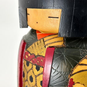 May include: A wooden doll with black hair, a red and gold patterned kimono, and a green and gold patterned robe. The doll's face is painted with a smile and closed eyes.