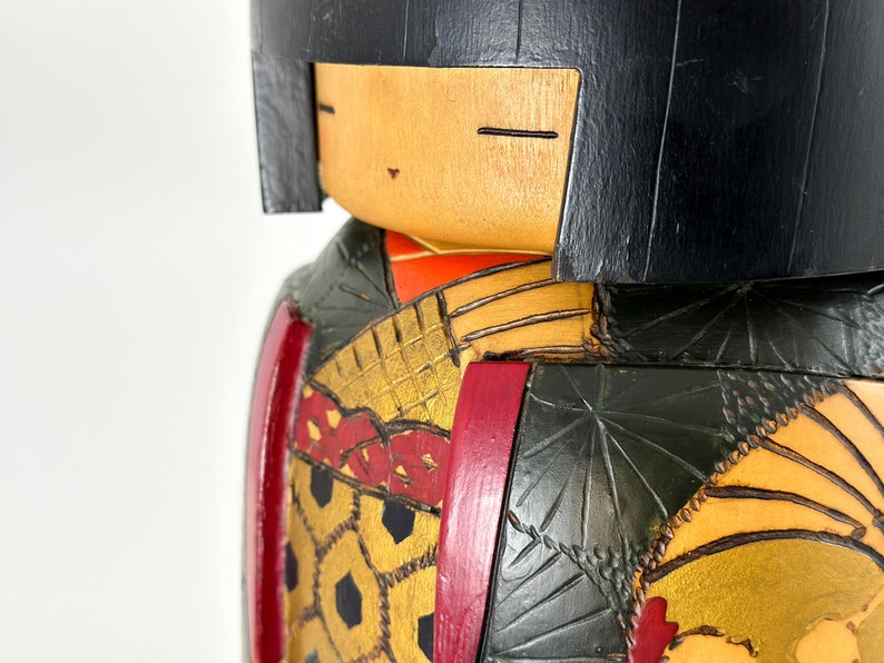 May include: A wooden doll with black hair, a red and gold patterned kimono, and a green and gold patterned robe. The doll's face is painted with a smile and closed eyes.