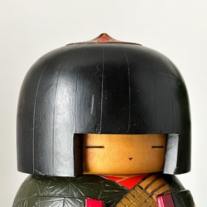 May include: A traditional Japanese wooden doll with a black hairpiece and a painted face. The doll is wearing a green and gold patterned kimono.