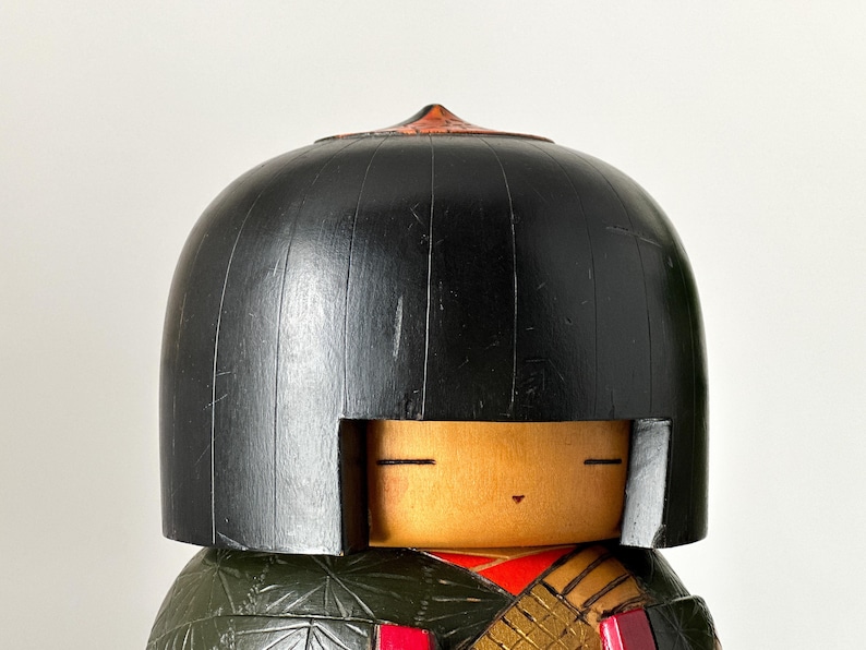 May include: A traditional Japanese wooden doll with a black hairpiece and a painted face. The doll is wearing a green and gold patterned kimono.