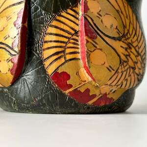 May include: A dark green wooden vase with a gold and red floral design. The vase has a round shape and a wide mouth.