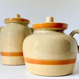 Op de afbeelding: Twee beige keramische potten met oranje banden en randen. Eén pot heeft een handvat, de andere heeft twee kleine handvatten. De potten hebben bijpassende deksels met oranje randen.