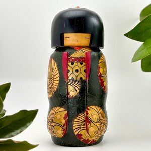 May include: A traditional Japanese kokeshi doll with a black hair, red and gold patterned kimono, and a black and gold background.