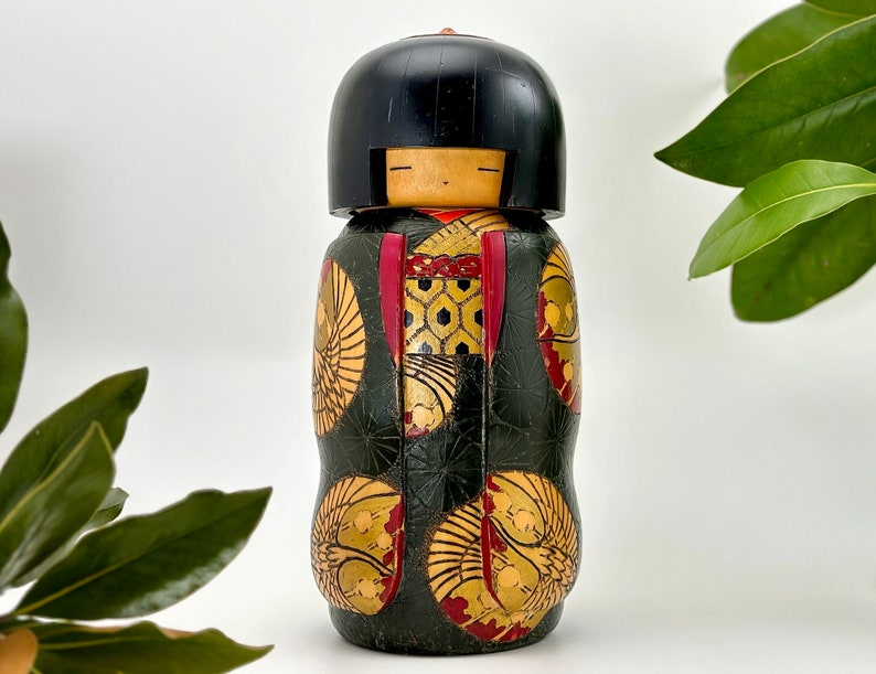 May include: A traditional Japanese kokeshi doll with a black hair, red and gold patterned kimono, and a black and gold background.