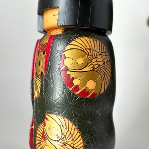 May include: A traditional Japanese kokeshi doll with a black hairpiece and a green, red, and gold patterned kimono. The doll is made of wood and has a round body.