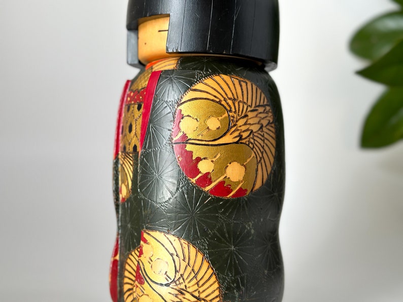 May include: A traditional Japanese kokeshi doll with a black hairpiece and a green, red, and gold patterned kimono. The doll is made of wood and has a round body.