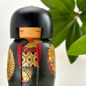 May include: A traditional Japanese kokeshi doll with black hair, a red and gold patterned kimono, and a black and gold background.
