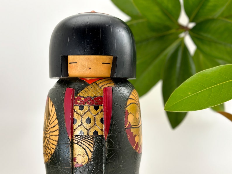 May include: A traditional Japanese kokeshi doll with black hair, a red and gold patterned kimono, and a black and gold background.