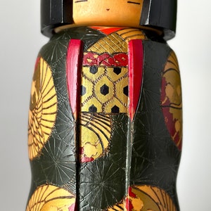 May include: A traditional Japanese kokeshi doll with a green, gold, and red patterned kimono. The doll has a black hair and a painted face.