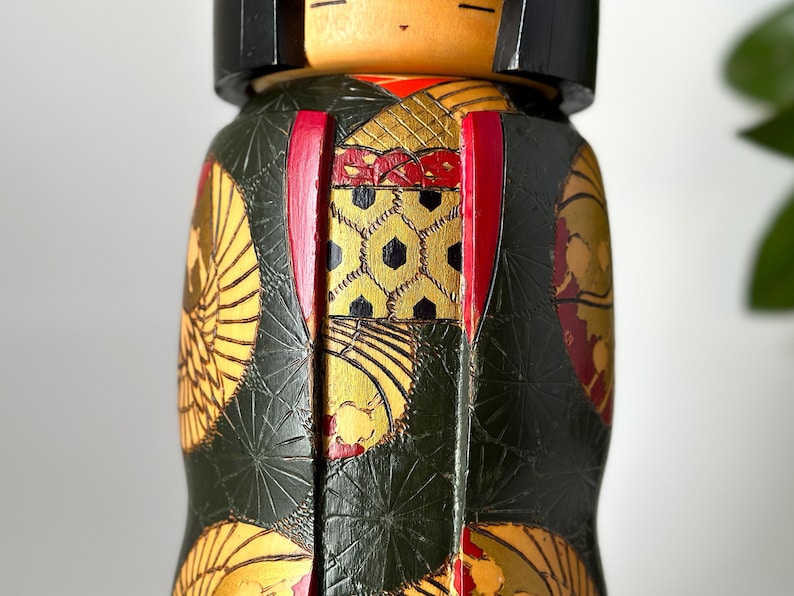 May include: A traditional Japanese kokeshi doll with a green, gold, and red patterned kimono. The doll has a black hair and a painted face.