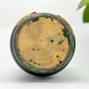 May include: The bottom of a wooden nesting doll, painted green with yellow and red accents. The wood is visible in the center of the doll.