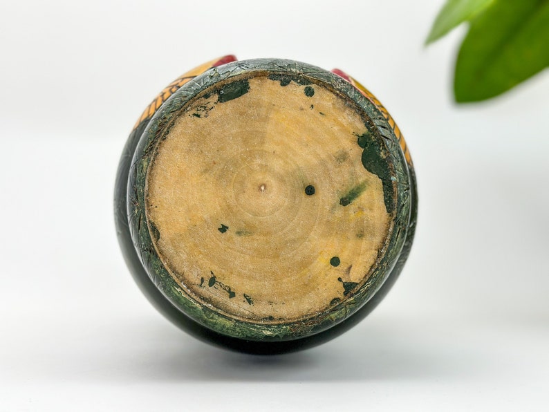 May include: The bottom of a wooden nesting doll, painted green with yellow and red accents. The wood is visible in the center of the doll.