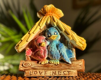 May include: A ceramic birdhouse with a thatched roof, featuring two birds inside. The blue bird is on the right and the pink bird is on the left. The birdhouse has a wooden base with the words "LOVE NEST" inscribed on it.
