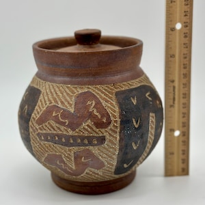 May include: A small, brown ceramic jar with a lid. The jar has a geometric design with brown, black, and white accents.