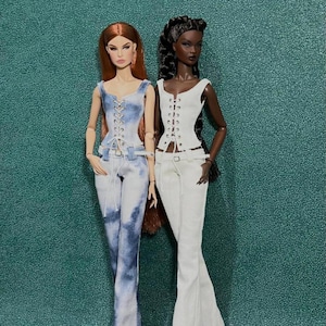 May include: Two fashion dolls wearing stylish outfits. One doll is in a denim-look corset top and matching jeans. The other doll is in a white corset top and white pants. Both dolls have long hair and are posed in front of a teal background.
