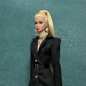 May include: A fashion doll with blonde hair in a high ponytail, wearing a black blazer-style dress with a belt. The doll is accessorized with gold earrings, a pearl and gem necklace, and a pendant. The doll has a full face of makeup.