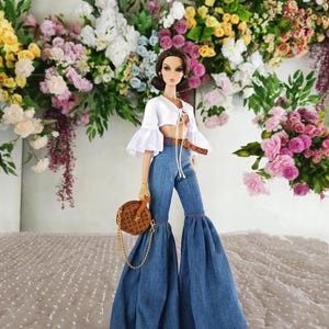 Op de afbeelding: Een modepop draagt een witte crop top met uitlopende mouwen, een blauwe spijkerbroek met wijde pijpen en een bruine riem. De pop draagt een bruine ronde handtas met een gouden ketting. Op de achtergrond een bloemstuk in diverse kleuren.