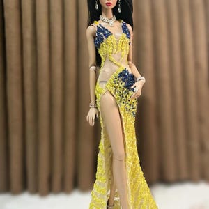 May include: A fashion doll wearing a yellow and blue beaded gown with a high slit. The doll has long black hair, red lipstick, and is accessorized with jewelry and silver heels. The dress features a sheer panel.
