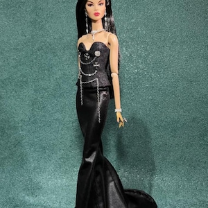 May include: A fashion doll in a black evening gown with a mermaid silhouette. The doll has long black hair and is adorned with silver jewelry, including a necklace, earrings, bracelet, and rings. The bodice features decorative chains and embellishments.