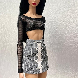 May include: A fashion doll with long black hair, wearing a black long-sleeved mesh crop top with black tape accents over the chest and a grey and white pinstriped mini skirt featuring a white lace-up front closure with silver metal eyelets.