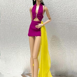 May include: A fashion doll with dark hair styled in a high ponytail, wearing a magenta halter-neck mini dress with a central cutout and a long, sheer yellow fabric drape attached to the side. The doll wears gold earrings and black high-heeled shoes.