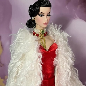 May include: A fashion doll with black hair styled in a bun, wearing a red velvet dress and a white, fluffy shawl. The doll is adorned with pearl earrings, a pearl and red bead necklace, and a gold chain. The doll's makeup includes dramatic eye shadow and lipstick.