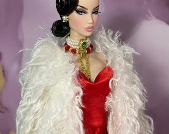 May include: A fashion doll with black hair styled in a bun, wearing a red velvet dress and a white, fluffy shawl. The doll is adorned with pearl earrings, a pearl and red bead necklace, and a gold chain. The doll's makeup includes dramatic eye shadow and lipstick.