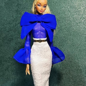 May include: A fashion doll wearing a royal blue top with a large bow detail and bell sleeves. The doll is also wearing a white lace pencil skirt and a gold belt. The doll has blonde hair and is wearing earrings.