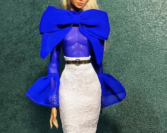May include: A fashion doll wearing a royal blue top with a large bow detail and bell sleeves. The doll is also wearing a white lace pencil skirt and a gold belt. The doll has blonde hair and is wearing earrings.