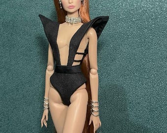 Può includere: Una bambola alla moda con lunghi capelli rossi, indossa un body nero con scollatura profonda e dettagli sulle spalle. La bambola è accessoriata con un girocollo scintillante, orecchini e braccialetti d'argento. Lo sfondo è verde acqua strutturato.
