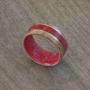 May include: A handcrafted ring with a vibrant red interior and a light-colored exterior band. The ring's design features a bold red center, framed by a lighter, possibly wooden, band. The ring is a unique piece of jewelry.