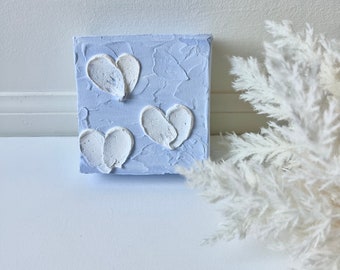 May include: A small, square canvas with a textured, light blue background. Three white, heart-shaped designs are raised from the surface. The artwork is displayed near a cluster of white, feathery decorative elements.