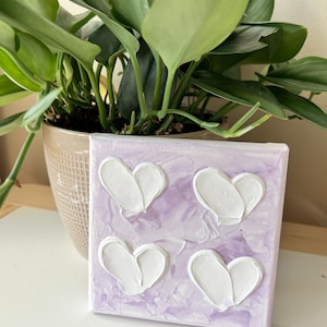 May include: A small canvas painting with a lavender background and four white raised hearts.