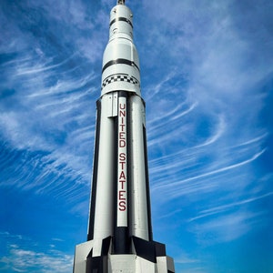 Puede incluir: Un cohete blanco y negro con las palabras "UNITED STATES" impresas en el lateral. El cohete está situado contra un cielo azul con nubes blancas.