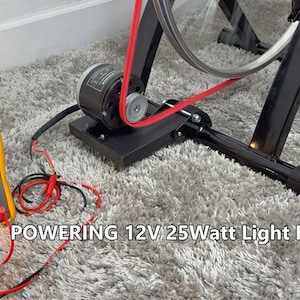 Puede incluir: Una bombilla encendida está conectada a un motor negro y a una rueda de bicicleta mediante cables rojos y negros. Un multímetro amarillo muestra una lectura. El texto "POWERING 12V 25Watt Light Bulb" es visible.