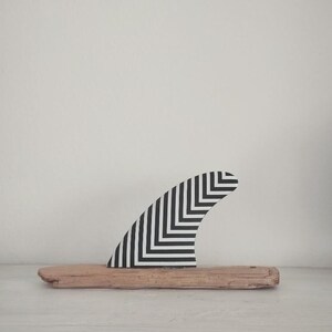 May include: A surfboard fin featuring a black and white chevron pattern mounted on a piece of light brown driftwood. The fin has a curved shape and is positioned upright on the horizontal piece of weathered wood, displaying a geometric design.