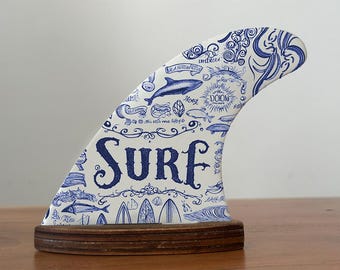 May include: A decorative surfboard fin with a white background and blue illustrations of marine life, waves, and the word "SURF". The fin is mounted on a wooden base, creating a unique coastal decor piece.