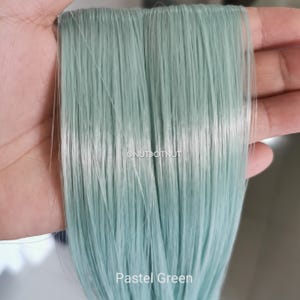 May include: A bundle of straight, synthetic hair extensions in a light pastel green color. The strands are smooth and uniform, featuring the text @NUTDOTNUT and the label Pastel Green printed clearly on the lower portion of the hair fibers.