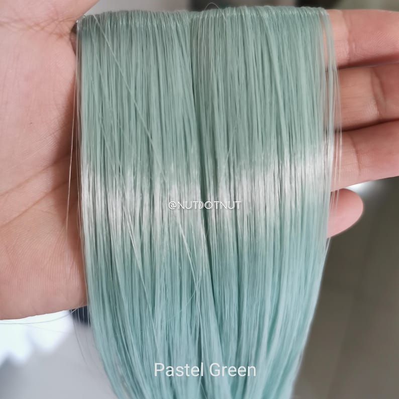 May include: A bundle of straight, synthetic hair extensions in a light pastel green color. The strands are smooth and uniform, featuring the text @NUTDOTNUT and the label Pastel Green printed clearly on the lower portion of the hair fibers.