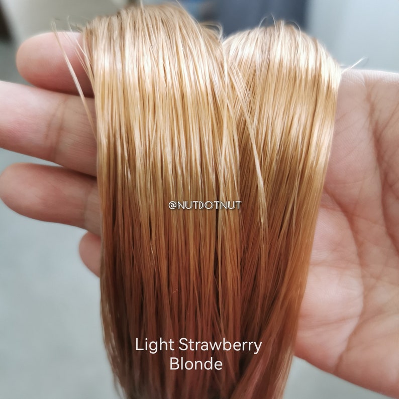 May include: A close-up of light strawberry blonde hair extensions. The hair is held in a hand and is very long and straight. The hair is a light blonde colour with a hint of pink.