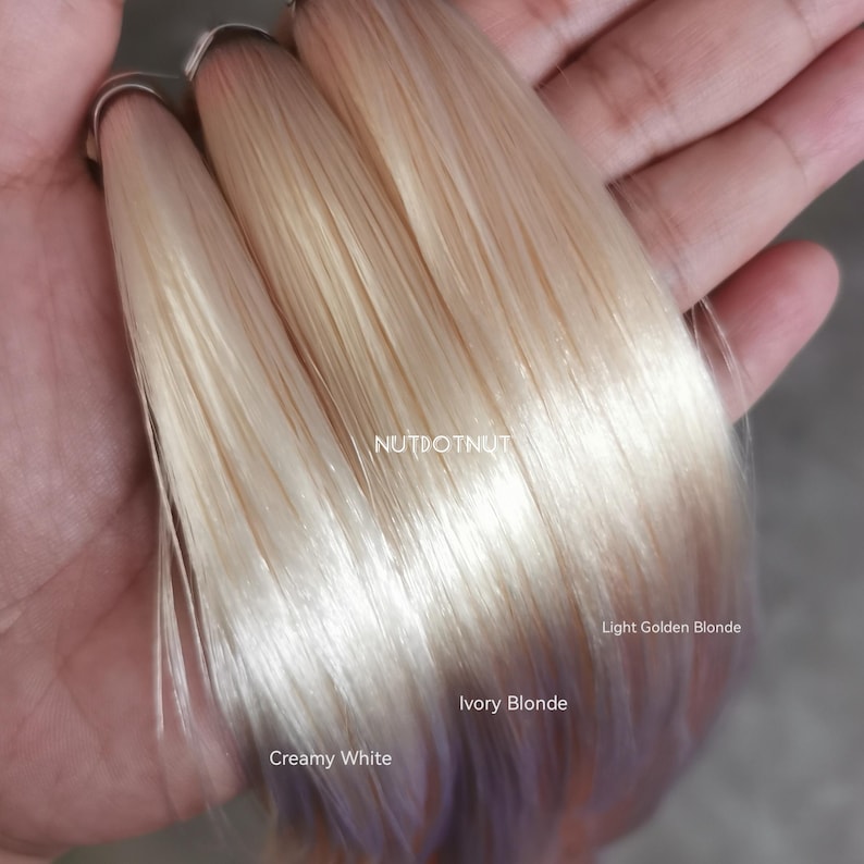 May include: Three strands of straight hair extensions arranged side by side, labeled with text. The colors are Creamy White, Ivory Blonde, and Light Golden Blonde. The brand name NUTDOTNUT is printed in the center of the hair strands.