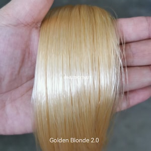May include: A straight, smooth lock of hair in a light golden blonde shade. The hair has a glossy, reflective finish and is presented in a uniform, vertical arrangement. The text "Golden Blonde 2.0" is printed at the bottom of the image.