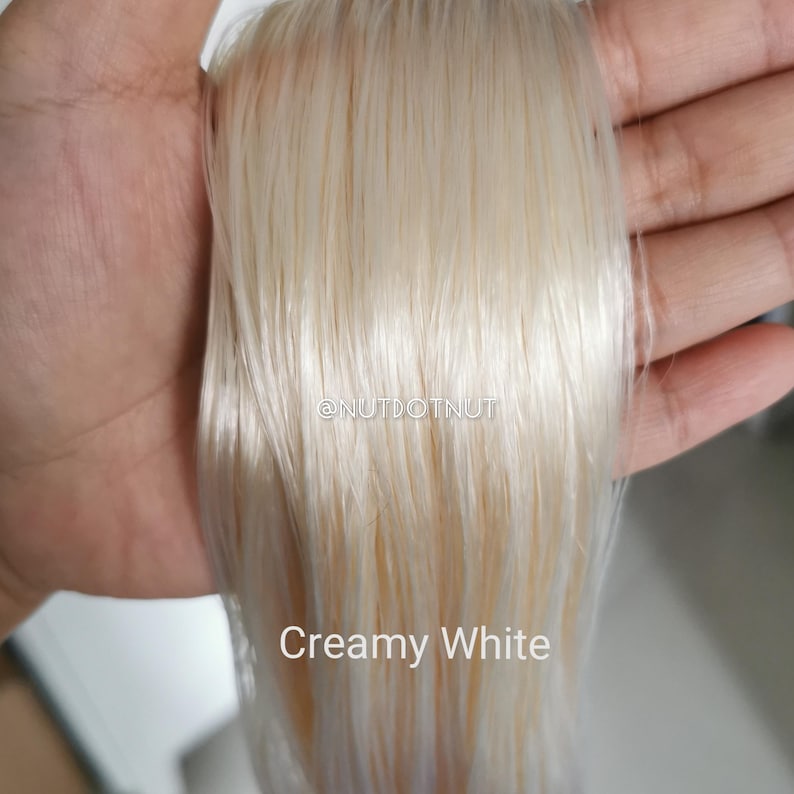 May include: A bundle of straight, smooth, creamy white synthetic hair extensions. The hair has a pale, off-white tone with subtle warm undertones. The text @NUTDOTNUT and the words Creamy White are printed in white on the hair strands.